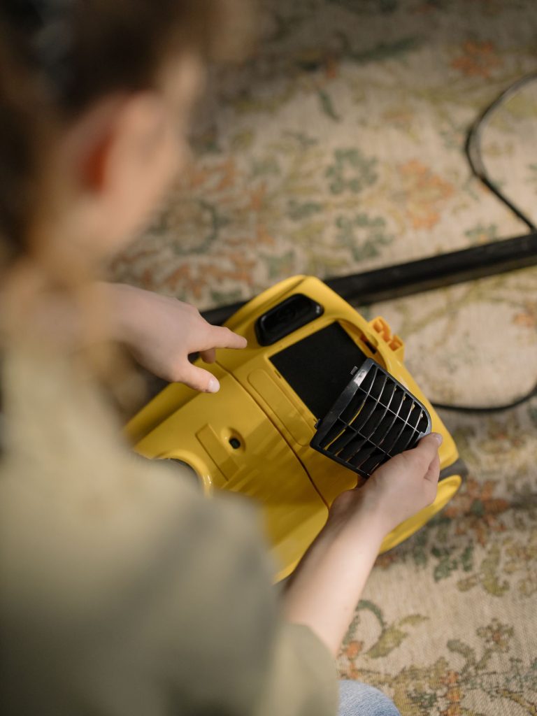 Hands servicing a yellow vacuum cleaner on a patterned carpet